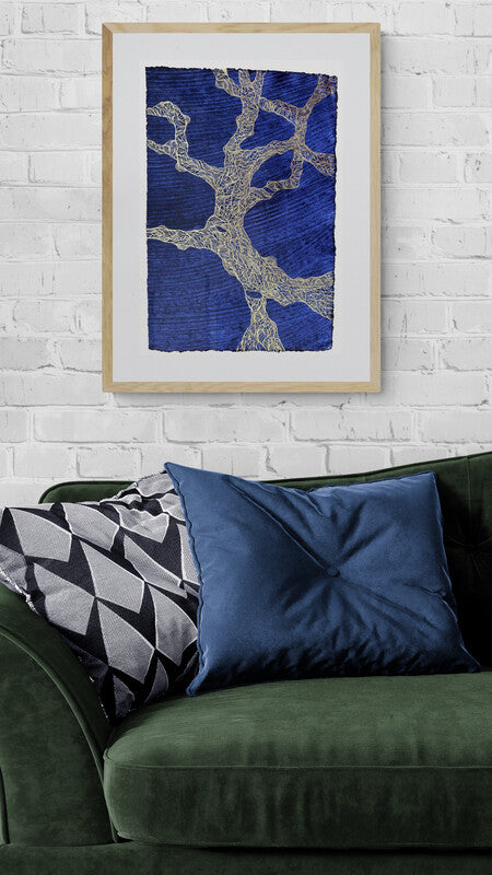 Drawing inspired by gnarly tree trunks and branches on indigo blue paper, framed and hanging on a white brick wall above a green sofa with a blue cushion.